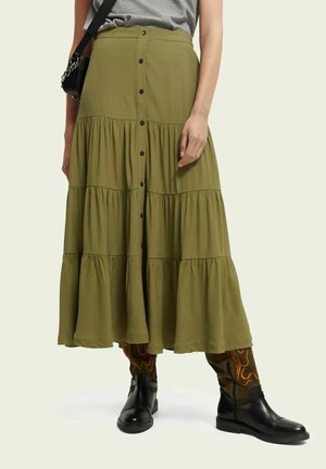 Olivgrüne, gestufte Midirock mit Knöpfen auf der Vorderseite. Leichter Stoff mit fließendem Design. Getragen mit schwarzen Stiefeln und gemusterten Hosen.