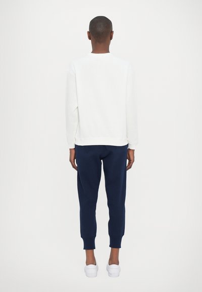Personne de dos portant une chemise blanche à manches longues, un pantalon de jogging bleu marine et des baskets blanches sur un fond blanc uni.