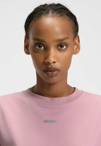 Giovane con capelli intrecciati che indossa una maglietta rosa con colletto rotondo con il logo "HUGO" centrato sul petto, che guarda dritto davanti a sé.