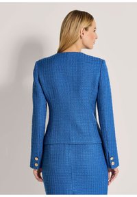 Blauwe textuur blazer met een gestructureerd ontwerp, lange mouwen en gouden knoopaccenten op de manchetten. Achteraanzicht met een getailleerde silhouet.