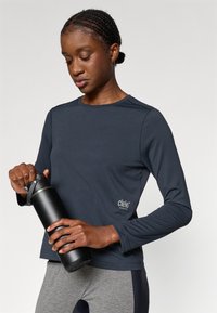 Dlhý rukáv športová blúzka v námorníckej modrej s logom na ľavej strane, skombinovaná s čiernou fľašou na vodu. Sivé textúrované legíny dotvárajú celkový vzhľad.