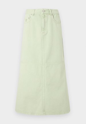 Femme debout portant une chemise blanche surdimensionnée, un haut marron, une jupe midi vert clair, des ballerines foncées, tenant un petit sac à main vert sur un fond uni.