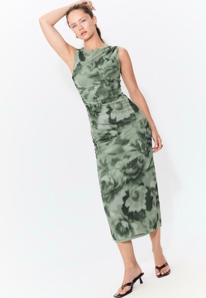 Grün floral bedrucktes Midi-Kleid mit figurbetonter Silhouette, gerafften Details und ärmellosem Design, kombiniert mit schwarzen offenen Mules.