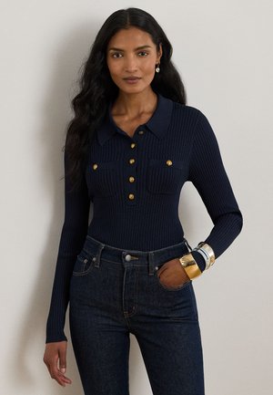 Femme aux longs cheveux foncés portant un haut côtelé bleu marine à manches longues avec des boutons dorés et un jean noir taille haute, accessoirisée avec des bracelets et des boucles d’oreilles.