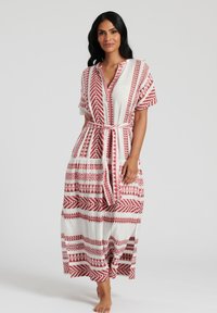 Maxiklänning i vitt tyg med röda geometriska mönster, korta ärmar och en knytbelte fram. Har en avslappnad passform och en flerdelad design.