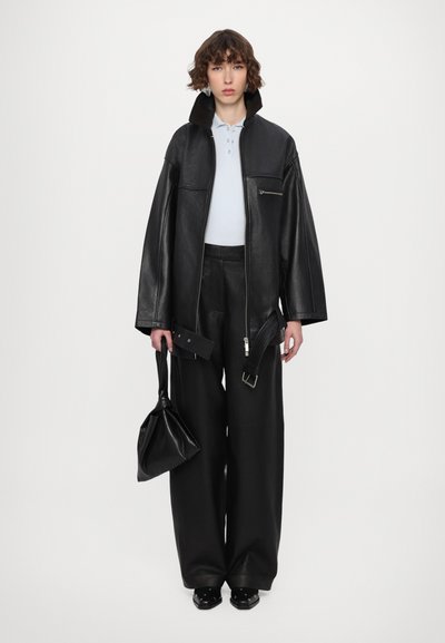 Must kombineeritud must nahast ülerõivastus kõrge kraega, sinine polo särg, mustad nahkpüksid ja must kottkott. Sile tekstuur, minimalism.