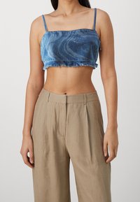 Top corto de mezclilla azul con dobladillo deshilachado y tirantes finos, que presenta un patrón en espiral y sutiles detalles de rhinestones. Llevado con pantalones beige.