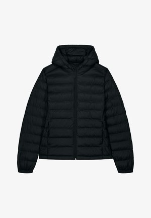 Femme debout les mains dans les poches, portant une doudoune noire, un jean gris ample et des baskets blanches sur un fond clair uni.