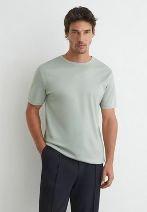Mand, der står op ad en hvid væg iført en lysegrå t-shirt med korte ærmer og mørke bukser, med den ene hånd i lommen.