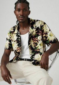 Camisa negra de flores de manga corta con grandes flores blancas y verdes, llevada sobre una camiseta blanca, combinada con pantalones beige.