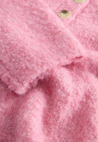 Rožinė megzta džemperis su minkštu, tekstūruotu paviršiumi. Pasižymi trimis arti išdėstytais apvaliais mygtukais ir laisvu, šiek tiek pūkuotu audiniu.