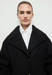Abrigo negro de gran tamaño con un amplio cuello, que presenta grandes botones. Debajo, se puede ver una camisa blanca con cuello. Tejido texturizado.