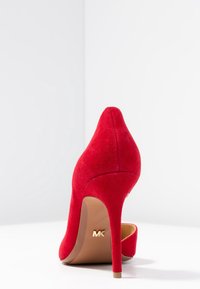 Escarpin en suède rouge à talon aiguille fin. Présente une pointe arrondie et un logo doré sur la semelle. Texture lisse.