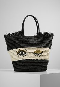 Sac tissé noir et crème avec deux motifs d'œil ; présente un paille texturé, un détail de frange en haut, et des poignées noires robustes.