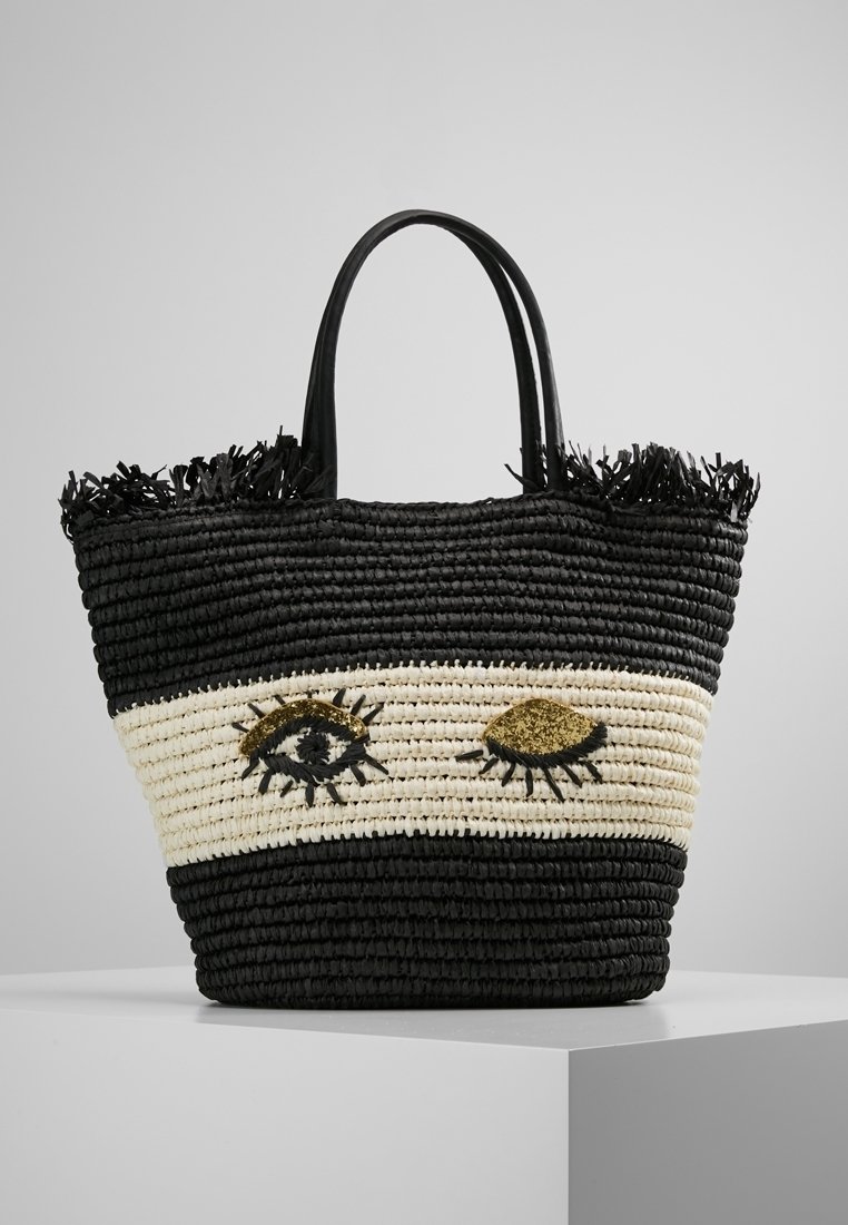 Sac tissé noir et crème avec deux motifs d'œil ; présente un paille texturé, un détail de frange en haut, et des poignées noires robustes.