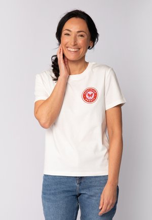 Frau mit dunklen Haaren im Pferdeschwanz, die lächelt, trägt ein weißes T-Shirt mit einem roten "Certified Beauty"-Logo und blaue Jeans, steht vor einem weißen Hintergrund.