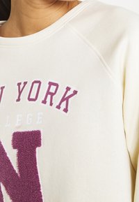 Primo piano di una felpa crema con una grande "N" viola testurizzata e il testo parziale "NEW YORK COLLEGE" in caratteri in stile varsity.