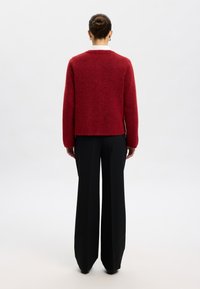 Rdeč volnen pulover s krojem v obliki škatle in zaobljenim izrezom, kombiniran s črnimi širokimi hlačami. Tkanina ima mehko, teksturirano površino.