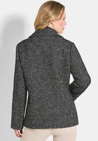 Vrouw met blond haar in een zwart en grijs herringbone patroon blazer, gezien vanuit de achterkant tegen een effen achtergrond.