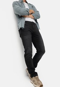 Black jeans worn with a light blue button-up shirt over a white T-shirt. Tan sneakers complete the outfit, highlighting casual style.