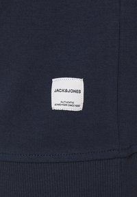 Mörkblå sweatshirttyg med en vit fyrkantig etikett som har "JACK&JONES" och "AUTHENTIC JEANSWEAR SINCE 1990." Ribbad detalj i nederkant.