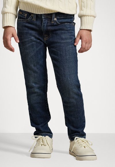 Pantalones vaqueros de mezclilla azul oscuro con un corte slim, sutiles desgastes y acentos de costura dorada, combinados con zapatos de lona beige claro.