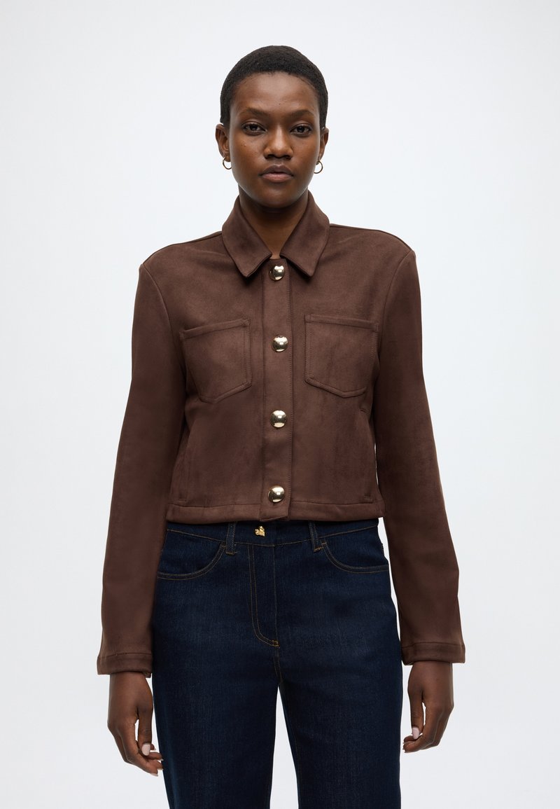 Braune Wildlederjacke in Kurzform mit Kragen, vorderen Taschen und großen Metallknöpfen. Kombiniert mit dunkelblauen Jeans. Glatte Textur.