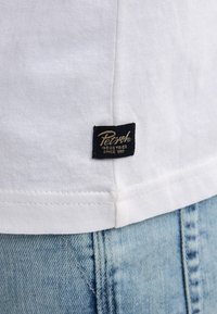 Der Saum eines weißen Baumwollhemds mit einem kleinen schwarzen Etikett, auf dem "Petrol Industries Since 1989" steht, über einer hellblauen Jeans.