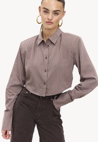 Chemise courte rayée brune avec un devant à boutons, un col pointu et des manches longues ; le tissu a une texture lisse et une coupe ajustée.