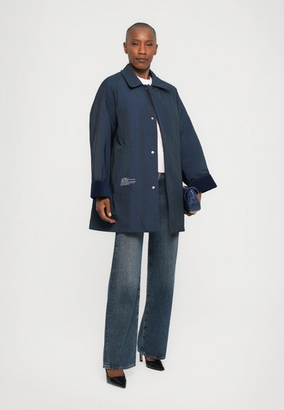 Navy blue oversized coat with a pointed collar, snap buttons, and wide cuffs. Paired with straight-leg denim and pointed black heels.