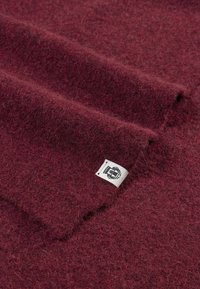 Bordeauxfarbener Wollstoff mit einer weichen, strukturierten Oberfläche, versehen mit einem kleinen Etikett mit einem in die Ecke gestickten Logo.