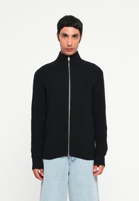 Svart ribbete zip-up cardigan med høy krage og lange ermer, med sølvglidelås og rett nederkant. Brukt over lyseblå jeans.