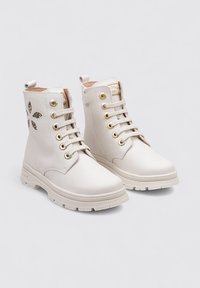 Bottes blanches à la cheville avec un laçage à l'avant, œillets dorés, languette au talon et design floral découpé sur le côté avec des détails métalliques.