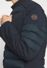 Marinhäftig jacka med en vadderad pufferdel på axeln, dragkedjestängning och en cirkulär logotyp på ärmen. Slät, texturerad tyg.