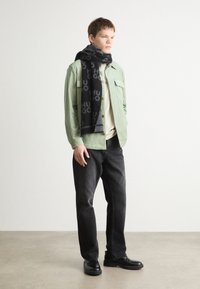 Fular negru din lână cu modelul logo-ului "HUGO", cămașă din flanel verde deschis, tricou bej, blugi negri și pantofi negri.