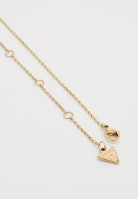 Gouden ketting met een delicate schakel, uitgerust met ronde schakels en een driehoekige hanger gegraveerd met "GUESS." Beveiligde musselklauwsluiting.
