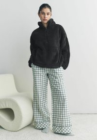 Zwarte fleece pullover met hoge kraag en rits, gecombineerd met broek in een breed geruit patroon in groen en wit, liggend op een zacht tapijt.