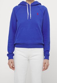 Blaues Baumwoll-Hoodie mit einer vorderen Tasche, weißen Kordeln und besticktem Logo auf der Brust, kombiniert mit weißen Hosen.