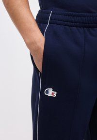 Το χέρι ενός ατόμου βρίσκεται στην τσέπη μπλε ναυτικού παντελονιού με λευκή ρίγα και ένα μικρό λογότυπο κροκόδειλου σε μπλε, λευκό και κόκκινο χρώμα.