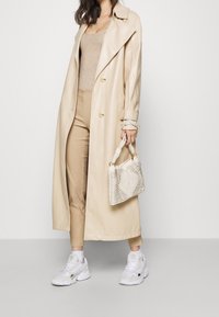 Beige läder trenchcoat med stora slag, ribbad beige tanktop, sandfärgade byxor och vita sneakers. Har en texturerad vit handväska.