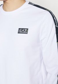 Vit långärmad skjorta med svartvitt mönstrat axelband och en liten svart EA7-logotyp på bröstet. Slät bomullstukt.