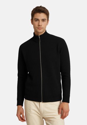 Jeune homme portant un pull noir zippé avec col et poignets côtelés, associé à un pantalon beige clair, debout devant un fond uni.