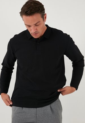 Camisa polo negra de manga larga con un tejido texturizado, tapeta de dos botones y cuello acanalado, utilizada sobre pantalones grises.