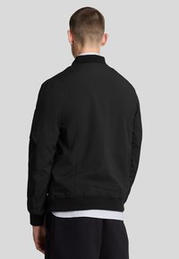 Giubbotto bomber nero con polsini e orlo a coste, caratterizzato da una tasca laterale e una texture liscia. Design semplice con una vestibilità classica.