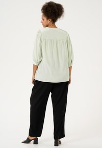Blusa verde chiaro, con texture, maniche a tre quarti, dettaglio arricciato sul retro e bottoni decorativi; abbinata a pantaloni neri svasati e sandali con tacco.