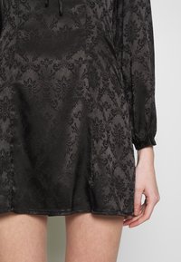 Černé šaty s texturovaným damaskovým vzorem, dlouhými rukávy se staženými manžetami a krátkou délkou. Hladký saténový materiál.