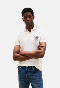 Polo de manga corta en color crema con cuello, detalles en azul marino y logotipo bordado en el pecho. Llevado con jeans de mezclilla azules.