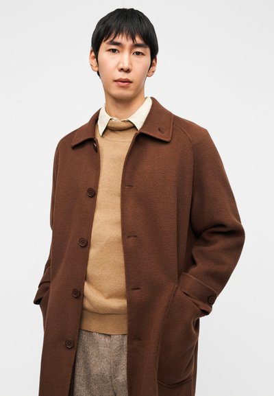 Καφέ παλτό από μείγμα μαλλιού με γιακά με σκίσιμο, με κουμπιά και πλαϊνές τσέπες, από πάνω ενός μπεζ πουλόβερ και υφασμένα παντελόνια.