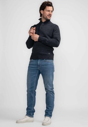 Donkergrijze trui met hoge kraag en knoopdetails, gecombineerd met lichtblauwe jeans en witte sneakers. De stof lijkt textuur te hebben.