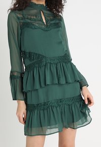 Mörkgrön klänning med långa ärmar, lager av volanger, spetsdetaljer och en nyckelhålsurklippning. Halvgenomskinligt tyg med texturerade detaljer.